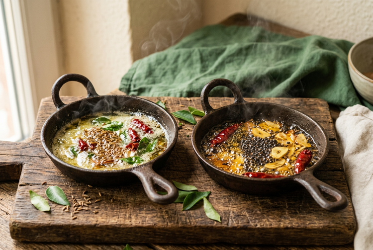 Two iron tadka pans side by side, one with ghee, cumin and curry leaves, the other with mustard oil, mustard seeds and garlic (illustrative image)